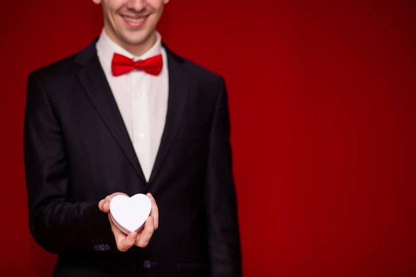 Καλαίσθητο άνθρωπος στο κοστούμι κρατώντας αγάπη δώρο κουτί με κόκκινο φόντο, εστίαση σε κουτί δώρου. Ημέρα του Αγίου Βαλεντίνου σύνθεση. Προσφορά γάμου - Φωτογραφία, εικόνα