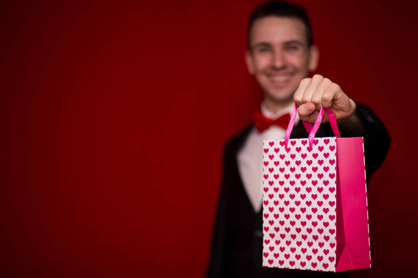 Young smiling man in suit hold love gift bag, with red background, focus on gift bag. Valentines day composition - Фото, зображення