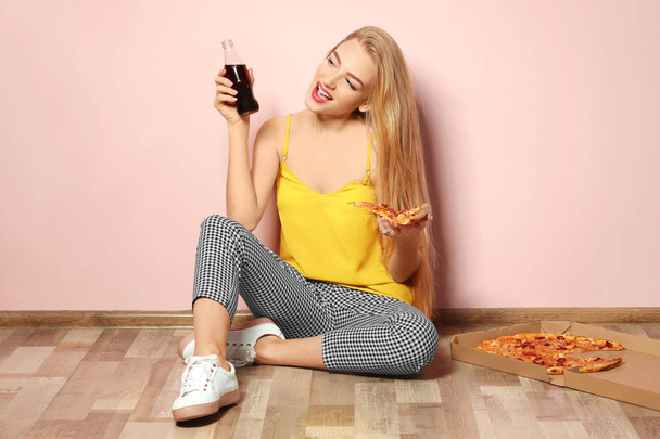 Attractive hipster girl with tasty pizza and bottle of cola near color wall - Valokuva, kuva