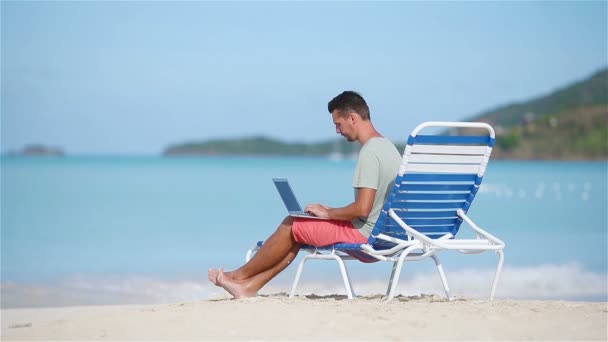 Młody człowiek z laptopa na tropikalnej plaży Karaibów. Człowiek siedzi na solarium z komputera i pracę na plaży - Materiał filmowy, wideo