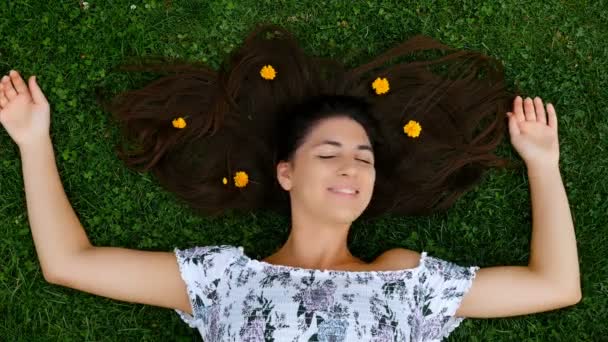 Young beautiful happy smiling girl lying on the grass with spreading hair, flowers falling over it. 4k - Footage, Video