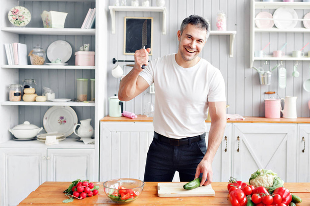 Портрет гарного усміхненого чоловіка на кухні. приготування їжі та концепція дому крупним планом чоловіча рука рубає огірок на обробній дошці з гострим ножем
 - Фото, зображення