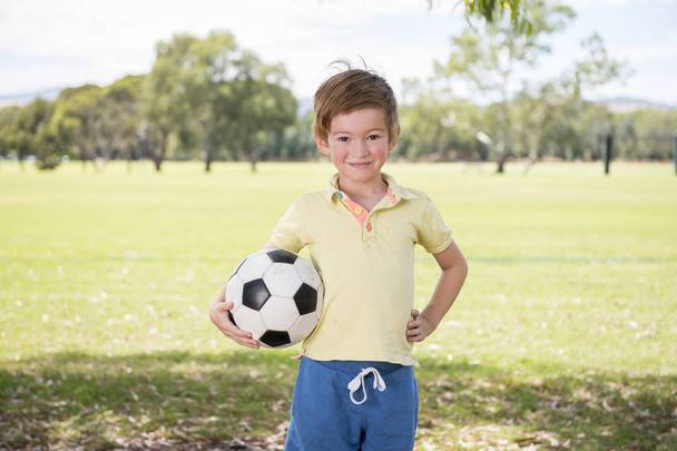 young little kid 7 or 8 years old enjoying happy playing football soccer at grass city park field posing smiling proud standing holding the ball  - Fotó, kép