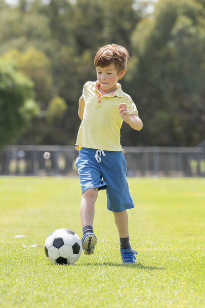 little kid 7 or 8 years old enjoying happy playing football soccer at grass city park field running and kicking the ball excited in childhood sport passion - Φωτογραφία, εικόνα