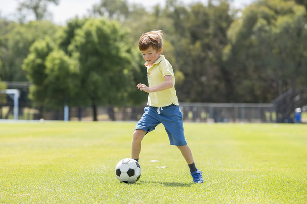 little kid 7 or 8 years old enjoying happy playing football soccer at grass city park field running and kicking the ball excited in childhood sport passion - Foto, Imagen