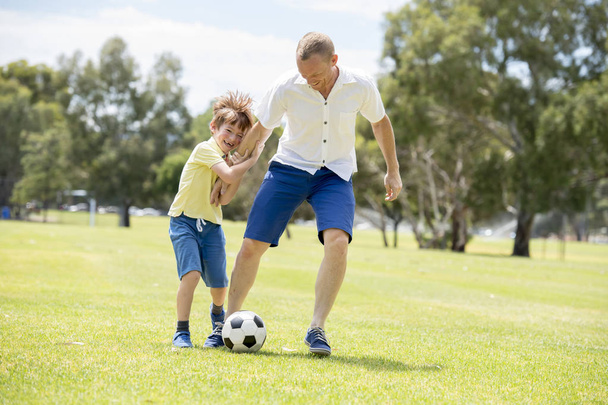 молодой счастливый отец и взволнованный маленький 7 или 8-летний сын играют вместе футбол в саду городского парка работает на траве ногами мяч
 - Фото, изображение