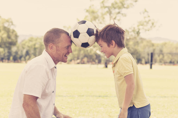 νέοι ευτυχής πατέρας και ενθουσιασμένος 7 ή 8 ετών ο γιος παίζει μαζί ποδόσφαιρο ποδόσφαιρο στο κήπο πάρκο πόλης θέτοντας γλυκό και αγαπώντας κρατώντας την μπάλα - Φωτογραφία, εικόνα