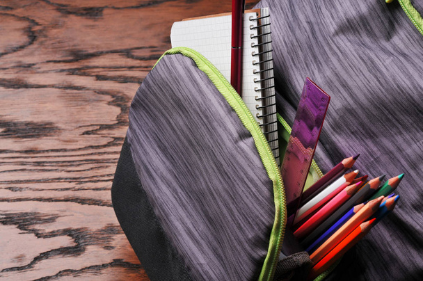 Close-up van volledige grijze school rugzak met kleur potloden, rode pen, liniaal en witte notebook op bruin houten tafel - Foto, afbeelding