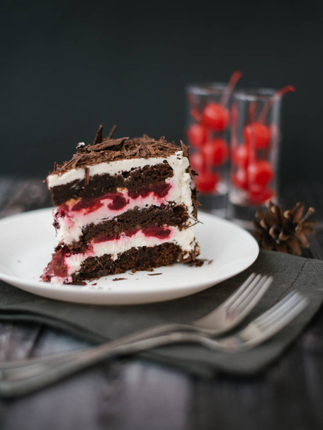 View of chocolate cake piece with cherries and yoghurt filling served on white plate with forks, napkin and pine cones on dark wooden table background - Zdjęcie, obraz