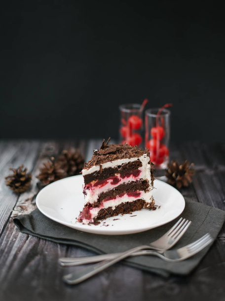 View of chocolate cake piece with cherries and yoghurt filling served on white plate with forks, napkin and pine cones on dark wooden table background - Foto, immagini