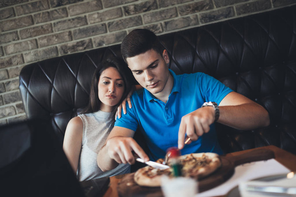 Atractiva y feliz pareja joven pasar un buen rato en el restaurante cafetería. Están sonriendo y comiendo una pizza - Foto, Imagen