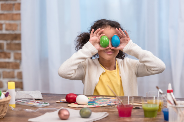 афроамериканська дитина розважається і прикриває очі фарбованими великодніми яйцями
 - Фото, зображення