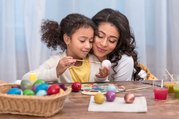 african american mother hugging daughter while she painting easter eggs - Фото, изображение