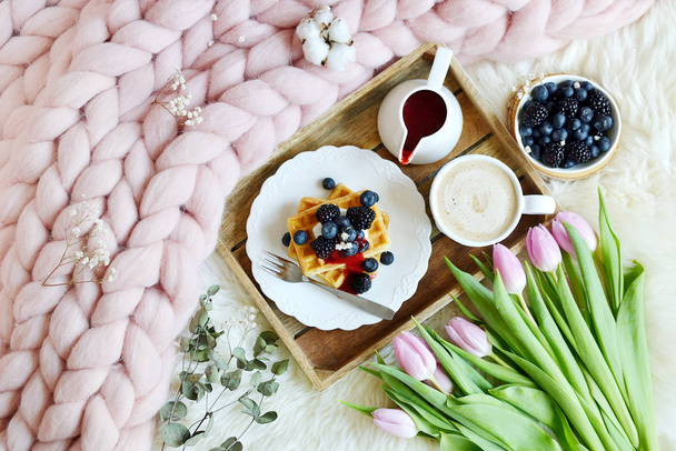 Cup met cappuccino en zelfgemaakte Belgische wafels met aardbei saus en bessen, roze pastel reus deken, bont, slaapkamer, roze bloemen tulpen, lente, vrouw dag ochtend concept  - Foto, afbeelding