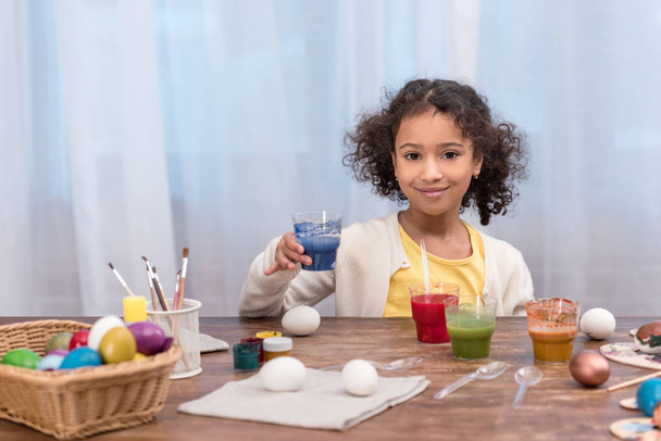 Afrikanisch-amerikanisches Kind hält Glas mit Farbe für Ostereier in der Hand und blickt in die Kamera - Foto, Bild