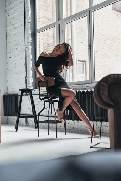 brunette woman in elegant black dress sitting on chair at home - 写真・画像
