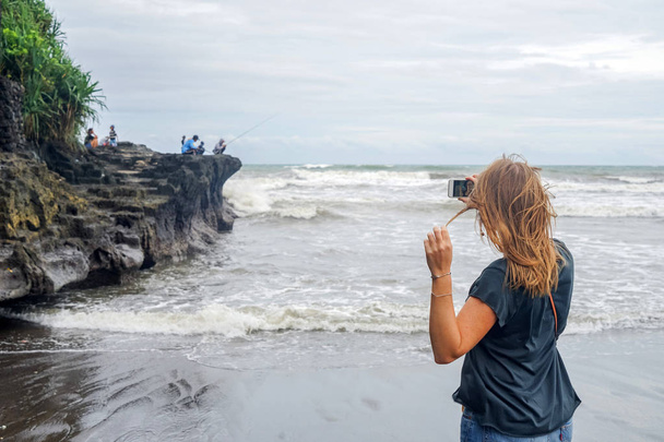 A young girl does selfie on a background of fishermen sitting on a rock and fishing in the ocean. Waves break on the rocks, a lot of splashes. The island of Bali. - Foto, Imagem