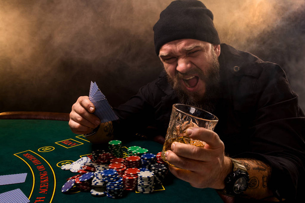Hombre barbudo con cigarro y vidrio sentado en la mesa de póquer en un casino. Apuestas, cartas y ruleta
. - Foto, Imagen