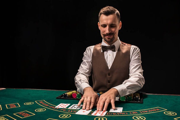 Portret krupierka trzyma karty do gry, hazard żetonów na stole. Czarne tło - Zdjęcie, obraz