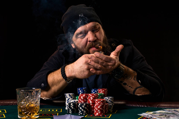Bebaarde man met sigaar en glas zitten aan een pokertafel in een casino. Gokken, speelkaarten en roulette. - Foto, afbeelding