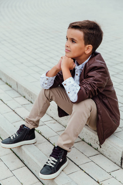 Adorable little boy sitting on stairs in a city, wearing a brown leather jacket. - Foto, imagen