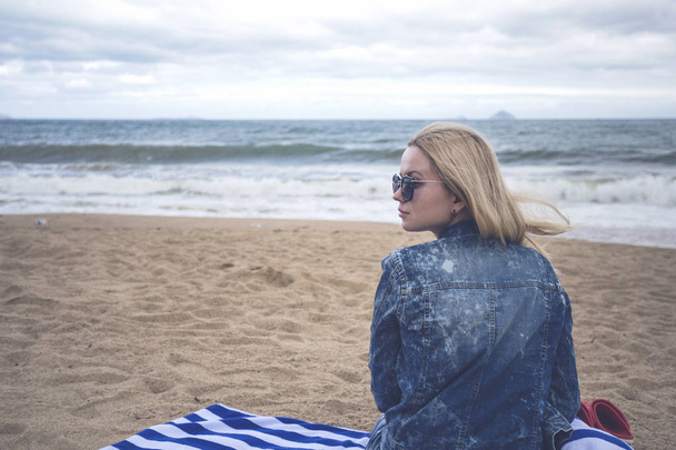 blonde girl in denim jacket and sunglasses sitting on the sea shore on a beach towel in a cold weather - Photo, Image