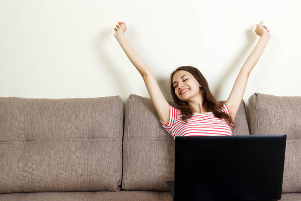 彼女のラップトップ コンピューターが付いているソファーに座っている若い女性は、空気、幸せと成功のコンセプトで手を挙げてください。 - 写真・画像