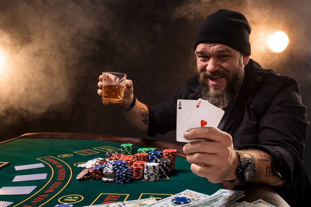 Мужчина играет в покер с сигарой и виски, мужчина показывает две карты в руке, выигрывая все фишки на столе с густым сигаретным дымом
. - Фото, изображение
