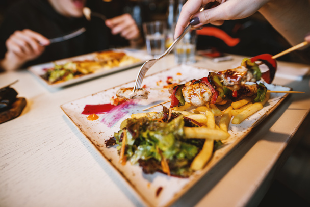 female hand eating fried skewers with chips and salad on plate in restaurant - Photo, Image