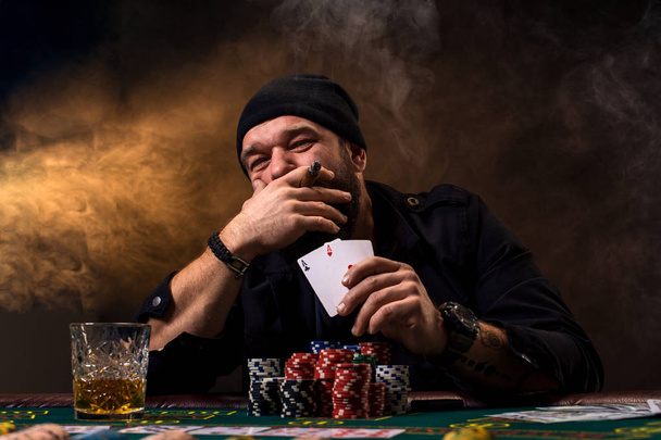 Man is playing poker with a cigar and a whiskey, a man show two cards in the hand, winning all the chips on the table with thick cigarette smoke. - Photo, Image