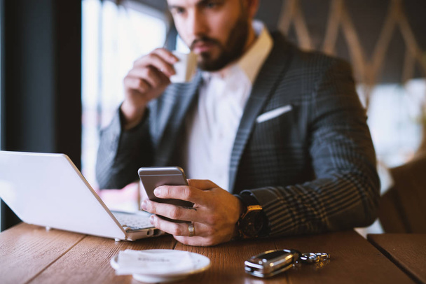 успешный красивый молодой бородатый бизнесмен в костюме работает с ноутбуком, пользуется смартфоном и пьет кофе в кафе
 - Фото, изображение