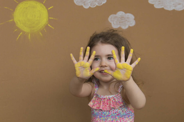 Käsitteellinen kesäloma. Söpö pieni tyttö uimapuku maalilla hänen kämmenet pelata taustalla maalattu aurinko ja valkoiset pilvet
. - Valokuva, kuva