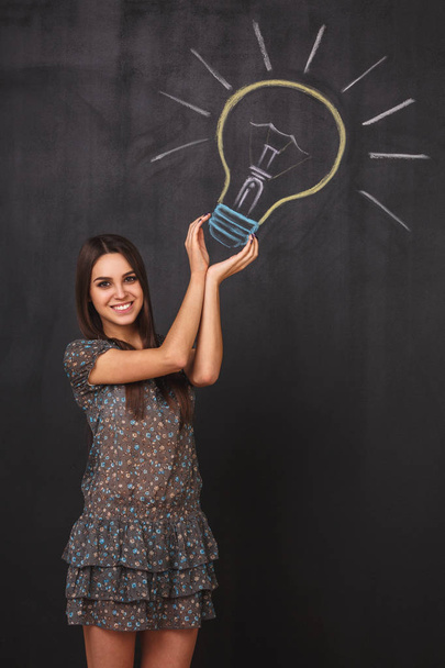 An attractive young girl has an idea. A happy student near the light bulb drawing on the blackboard. Catch the idea concept - Φωτογραφία, εικόνα