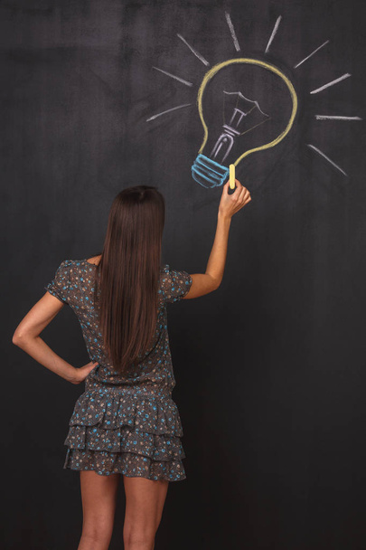 A young brunette girl draws multicolored light bulb on a black board. Light bulb with symbol business concept.  - Fotó, kép
