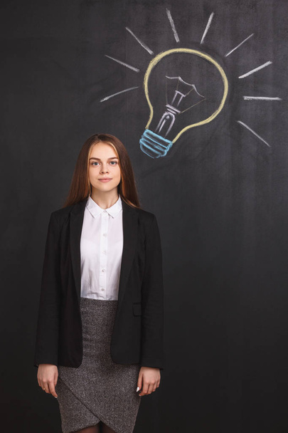 The successful young businesswoman has a great idea. The business lady near a colored lightbulb on the blackboard. - Foto, imagen