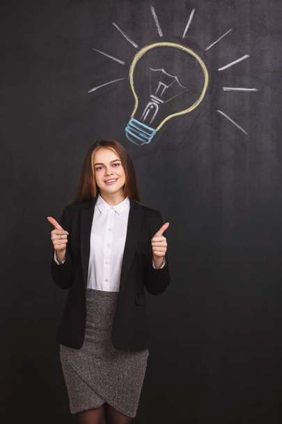 A young student near a lightbulb on blackboard. A business woman points upwards with thumbs up showing an approving gesture. - Foto, Bild