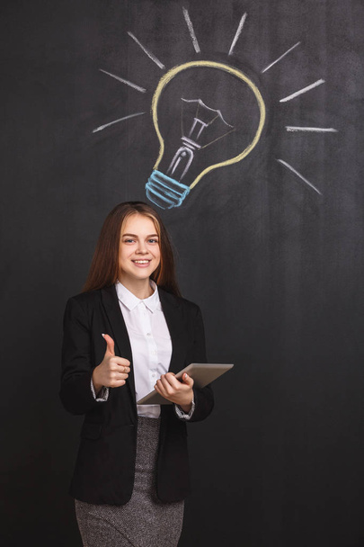 A young business woman points upwards with thumb up showing an approving gesture. A lady with a laptop near a lightbulb on blackboard. - Foto, Imagem