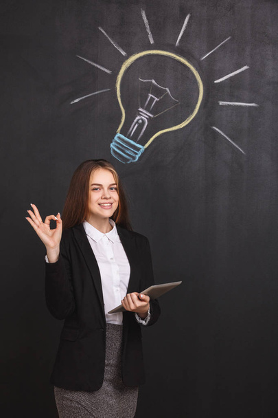 A young businesswoman with a laptop has an idea. A business lady with ok sign near a lightbulb on blackboard. - Photo, image