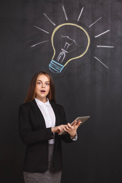 A business lady with open mouth near a lightbulb on the blackboard. The expressive young woman with a laptop has an idea - 写真・画像
