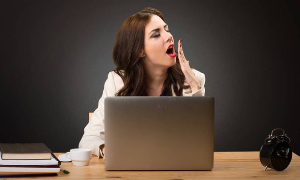 Mujer de negocios trabajando con su portátil y bostezando sobre fondo negro
 - Foto, Imagen