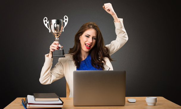 Business woman working with her laptop and holding a trophy on black background - Photo, Image