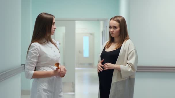 Μια γυναίκα γιατρό και μια νεαρή έγκυο γυναίκα σταθεί στο διάδρομο του κέντρου μεταμόσχευσης και ο γιατρός ρωτά ερωτήσεις σχετικά με την κατάσταση και την πορεία της εγκυμοσύνης. Ακουμπά το στομάχι μαζί της - Πλάνα, βίντεο