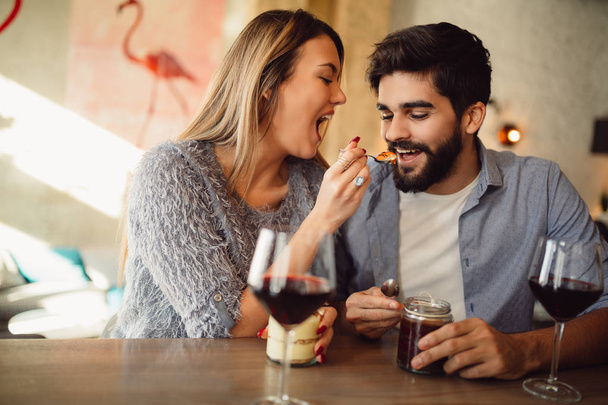 Красивая девушка кормит своего парня тортом в кафе, пьет вино и празднует День Святого Валентина
. - Фото, изображение