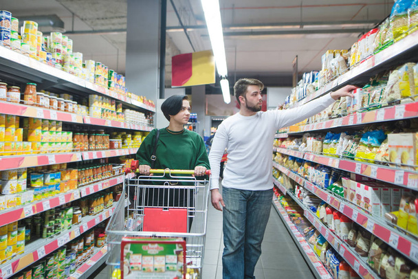 Una giovane bella coppia che cammina con un carrello del supermercato e sceglie i prodotti. Shopping in famiglia in un supermercato
 - Foto, immagini