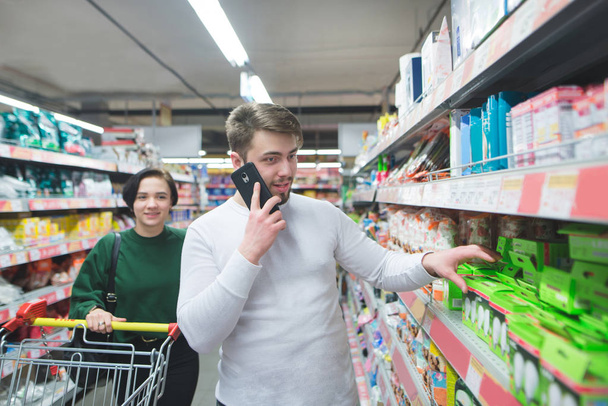 Ένας άνθρωπος μιλάει τηλεφωνικώς σε ένα σούπερ μάρκετ και επιλέγει αγαθά. Η κοπέλα πηγαίνει με ένα καλάθι του σούπερ μάρκετ. Ψώνια στο σούπερ μάρκετ - Φωτογραφία, εικόνα