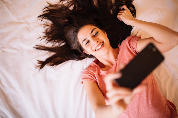 Mooie jonge meisje dat neemt selfie of video chatten terwijl liggend op het bed. Top hoekmening. - Foto, afbeelding