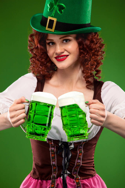 St Patricks Day. Young sexy Oktoberfest waitress, wearing a traditional Bavarian dress, serving big beer mugs on green background with copy space - Fotó, kép