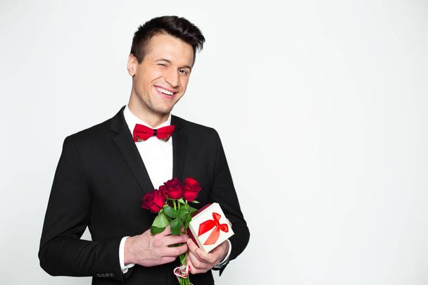 Stylish smiling handsome modern man in a official black suit and a red bow tie is holding a gift box and beautiful red roses in hands. Gift for beloved woman of friend. - Valokuva, kuva