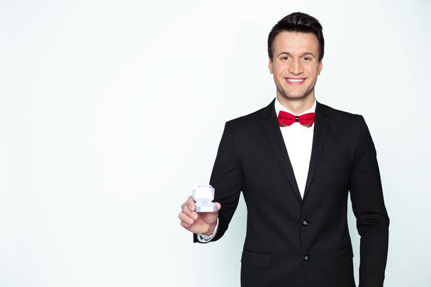 Pohledný usmívající se moderní elegantní muž v oficiální černý oblek a červený motýlek drží box s zásnubní prsten a ukazuje ve fotoaparátu. - Fotografie, Obrázek