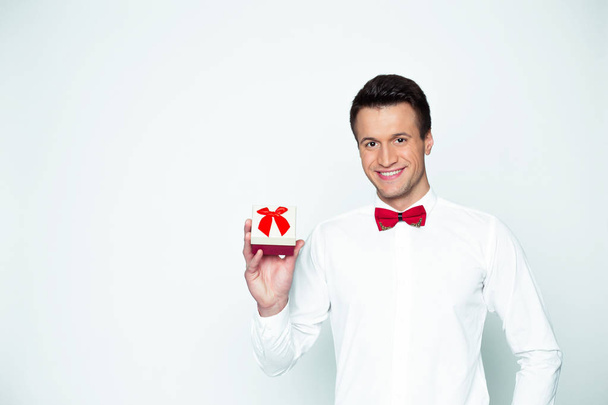 A gift for the beloved. Young beautiful smiling elegant man in a white shirt and red bow tie holding a box with a gift and shows it to the camera. Valentine's Day. - Photo, Image